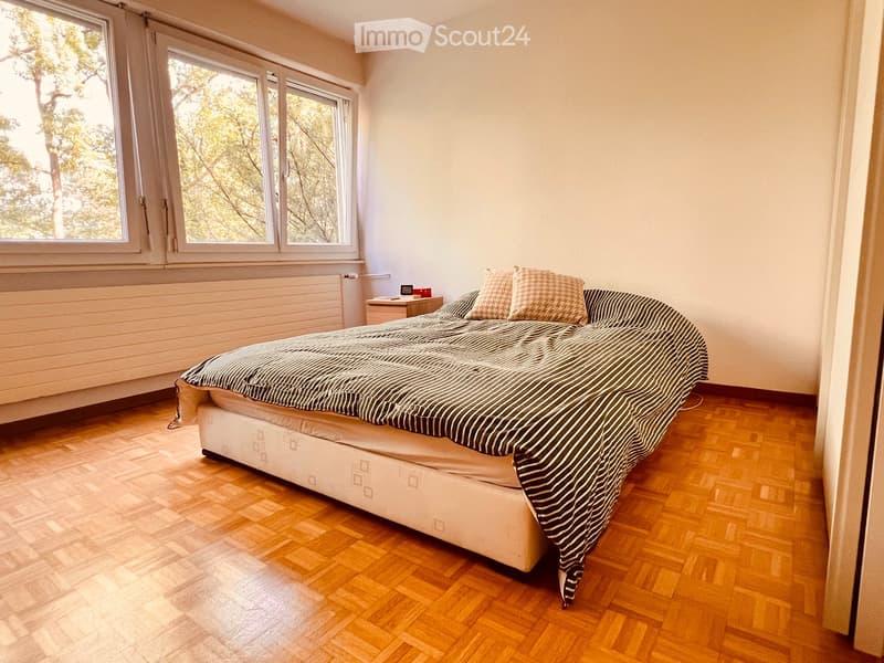 Bright bedroom with wooden flooring, large windows, a neatly made bed with striped bedding, a nightstand, and a radiator below the windows.