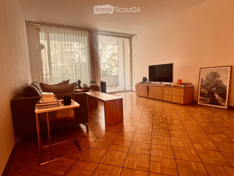 Living room with wooden flooring, large window with sheer curtains, balcony access, sofa, coffee table, bench, TV, and cabinet.
