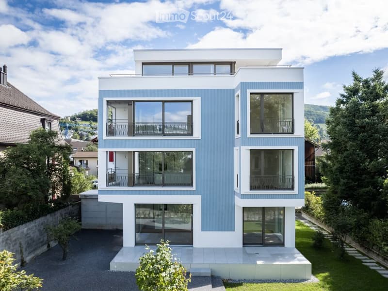 Modern two-story house with blue and white exterior, large windows, balconies, and a raised ground floor.