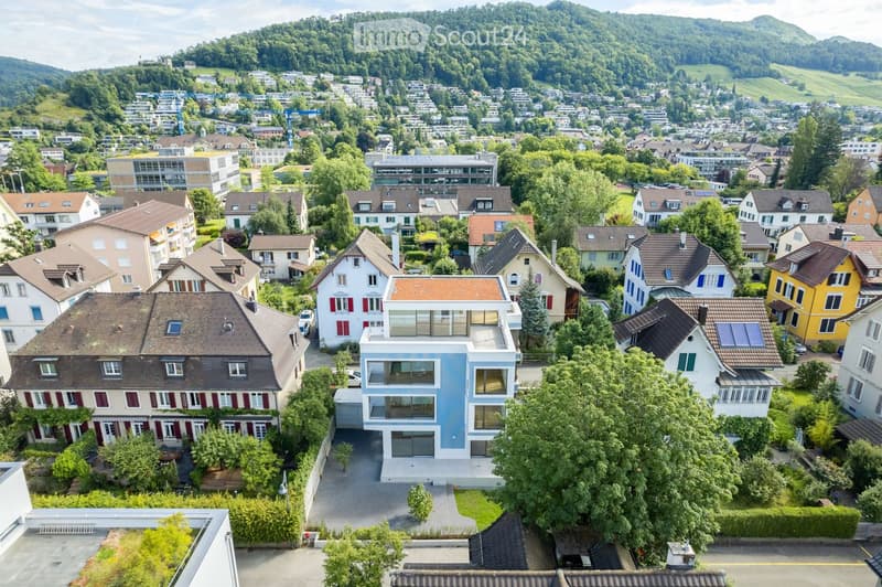 Aerial view of a residential neighborhood with various houses, trees, and a scenic backdrop of hills and greenery.
