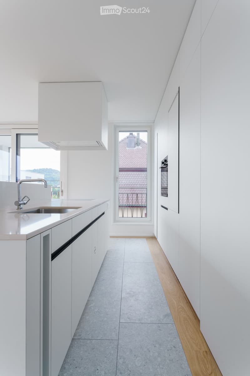 Modern, minimalist kitchen with white cabinets, a stainless steel sink, and a sleek faucet. The space features a large window offering a view of the roof and surrounding area.
