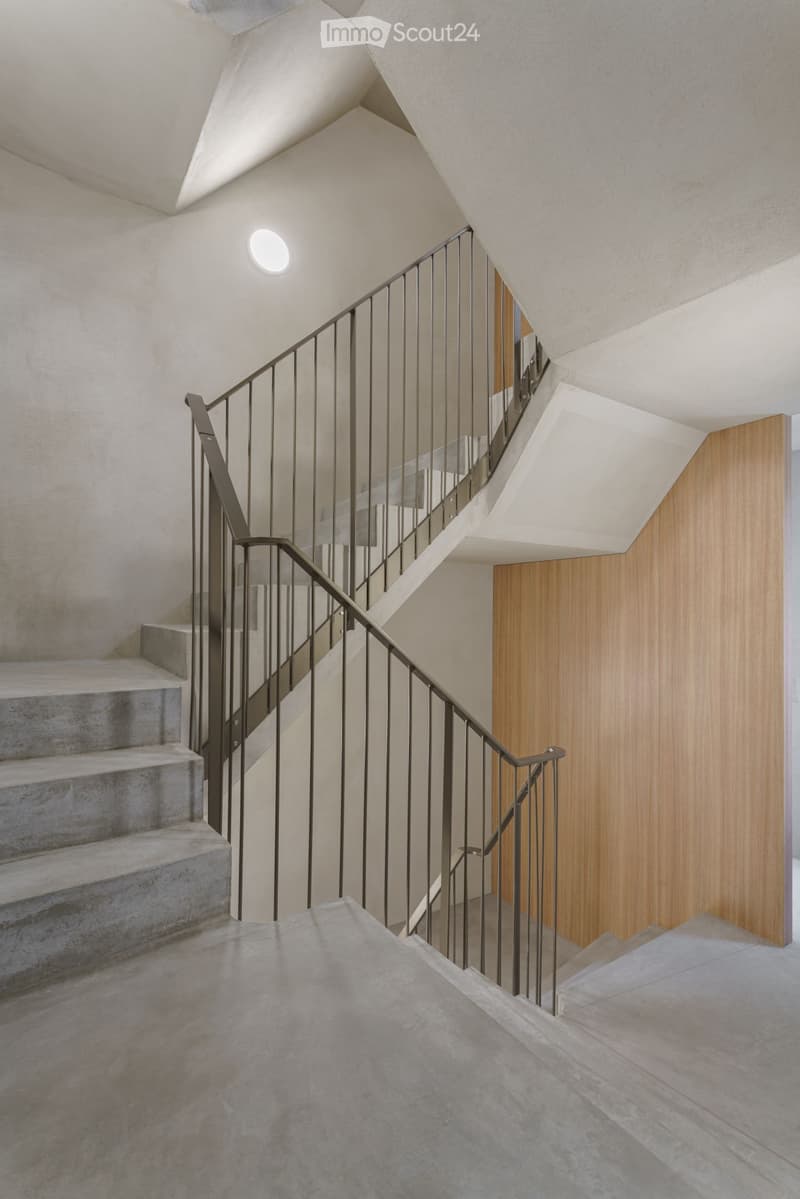 Modern staircase with concrete steps, sleek metal railing, and warm wooden paneling on one wall. The ceiling features recessed lighting and a geometric design.