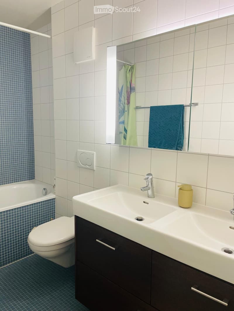 Modern bathroom with white tiles, a double sink, a toilet, a bathtub, and a large mirror. Blue towels are hanging on a rack, and a yellow soap dispenser is placed on the sink.