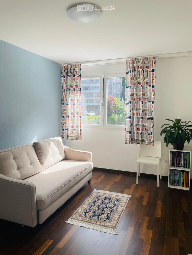 Living room with a light blue wall, wooden floor, and large windows with colorful curtains. Features a beige sofa, white chair, bookshelf with books, a potted plant, and a patterned rug.