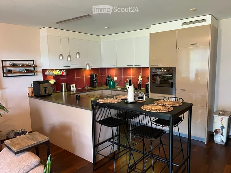 Modern kitchen with white cabinets, stainless steel appliances, and a black island with bar stools. Features include a wine rack, pendant lights, and a small seating area with a wooden bench.