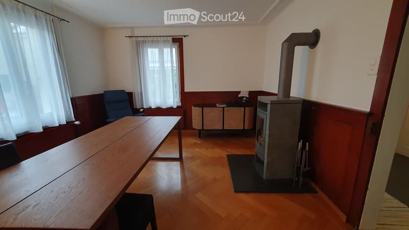 A room with wooden flooring and walls, featuring a long wooden table, a blue chair, a wooden stove, and a storage unit with wicker baskets.
