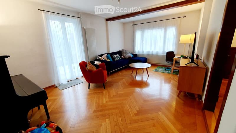 Living room with wooden floor, large windows, blue sofa, red armchair, coffee table, small table, and piano.