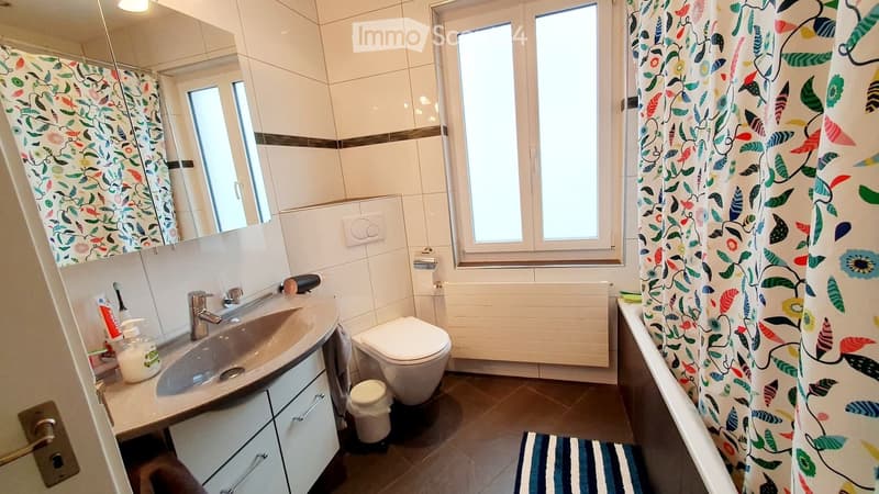 Modern bathroom with white tiles, a large window, a sink with a mirror, a toilet, and a shower curtain. Includes a soap dispenser, toothpaste, and a small trash bin.