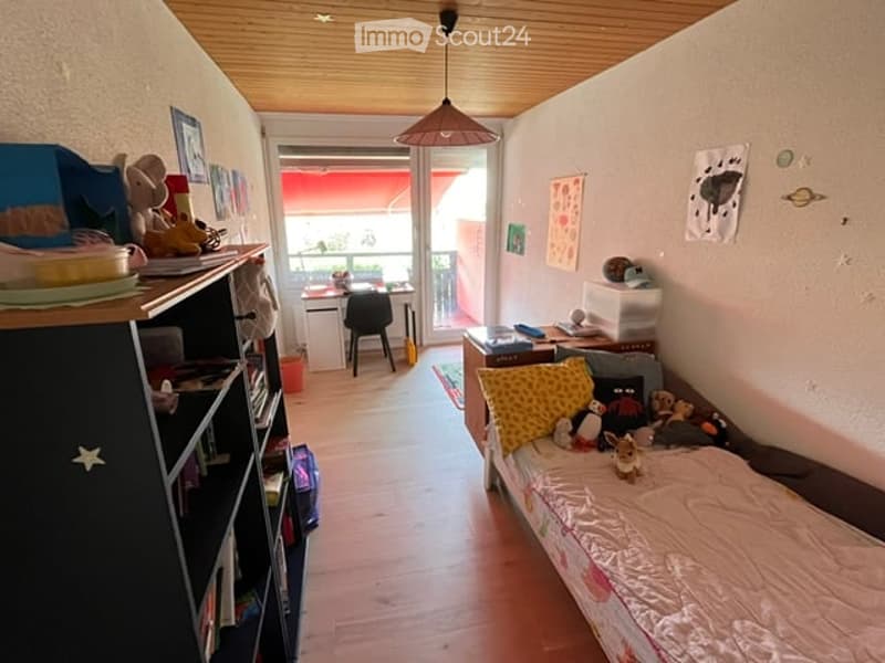 Cozy children's bedroom with a single bed, bookshelf, small table, and balcony access. The room features wooden flooring and a hanging light fixture.
