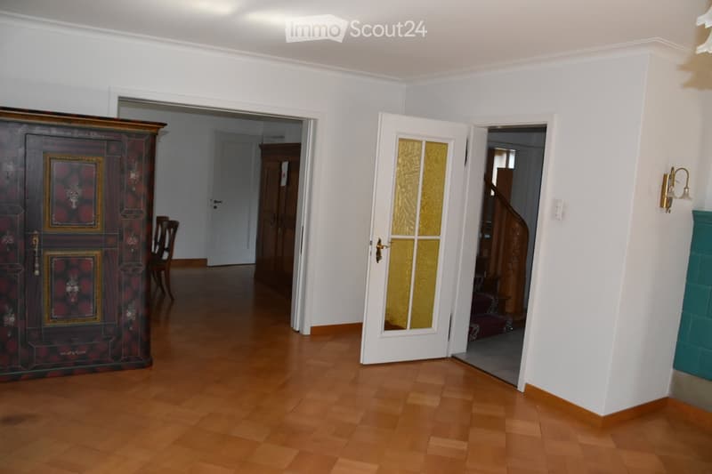 Bright hallway with wooden flooring, white walls, and open doors leading to other rooms. Features a decorative cabinet, a chair, and a staircase visible in the background.