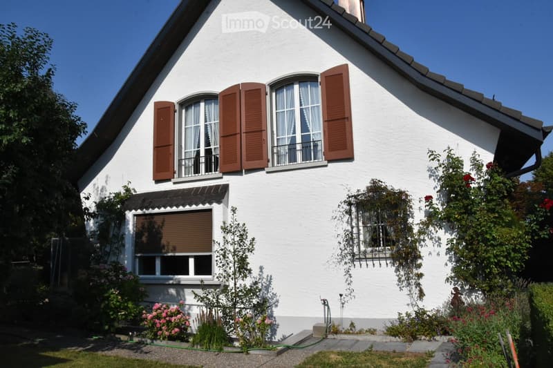 Two-story white house with brown shutters, multiple windows, and a small balcony. The house is surrounded by a garden with various plants and flowers.