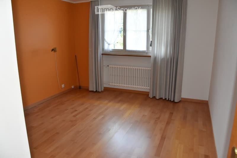 Empty room with wooden flooring, orange accent wall, large window with curtains, radiator, and a broom leaning against the wall.