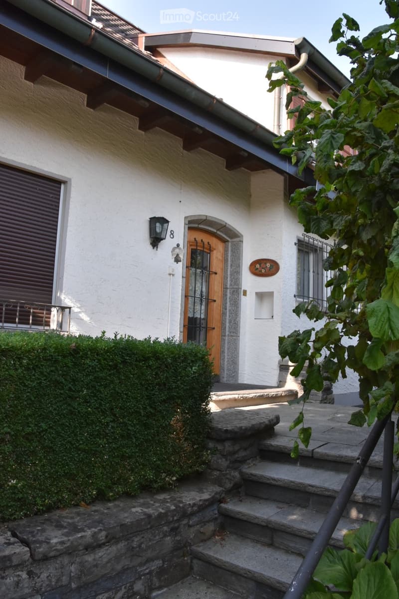 Two-story house with white exterior, wooden front door, stone steps, and greenery.