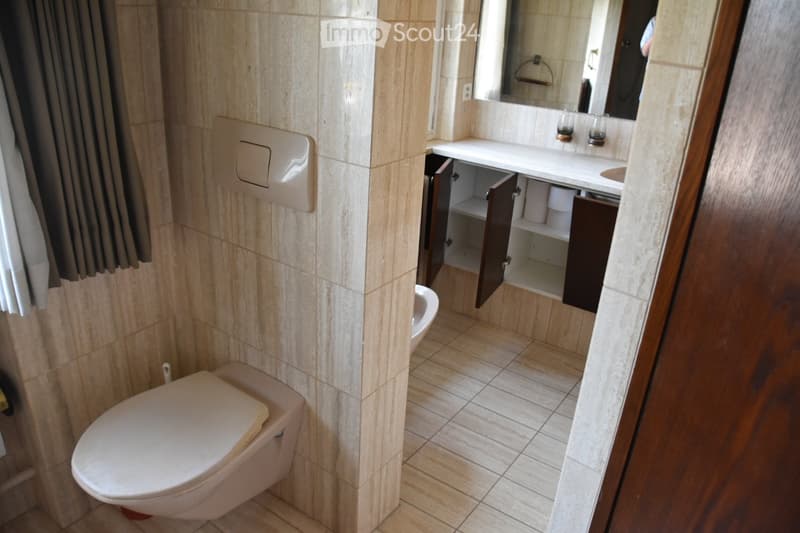 Modern bathroom with beige tiled walls and floor, a toilet, a sink with a mirror above it, and an open cabinet with shelves.