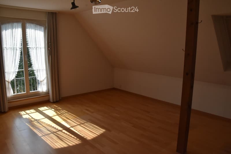 Empty attic room with wooden flooring, large window with white curtains, and slanted ceiling beams.