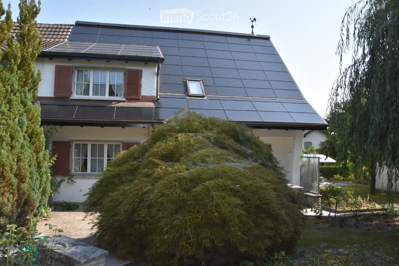 Two-story house with a modern, sloped roof featuring solar panels. The exterior is white with red shutters and multiple windows. The house is surrounded by greenery, including trees and shrubs, with a gravel driveway leading to the entrance.