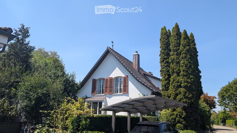 A two-story white house with a brown roof and shutters, surrounded by trees and a carport with a parked car.