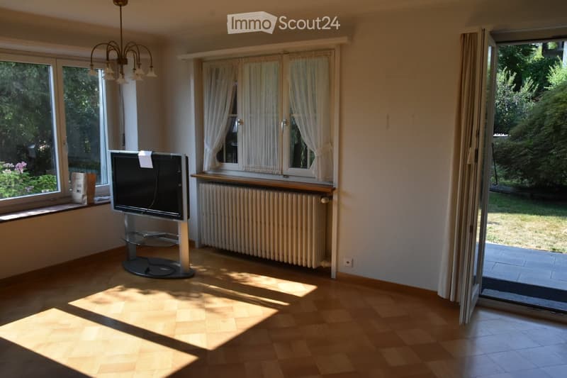 Bright living room with wooden flooring, large windows, a television on a stand, radiator, and a chandelier hanging from the ceiling.