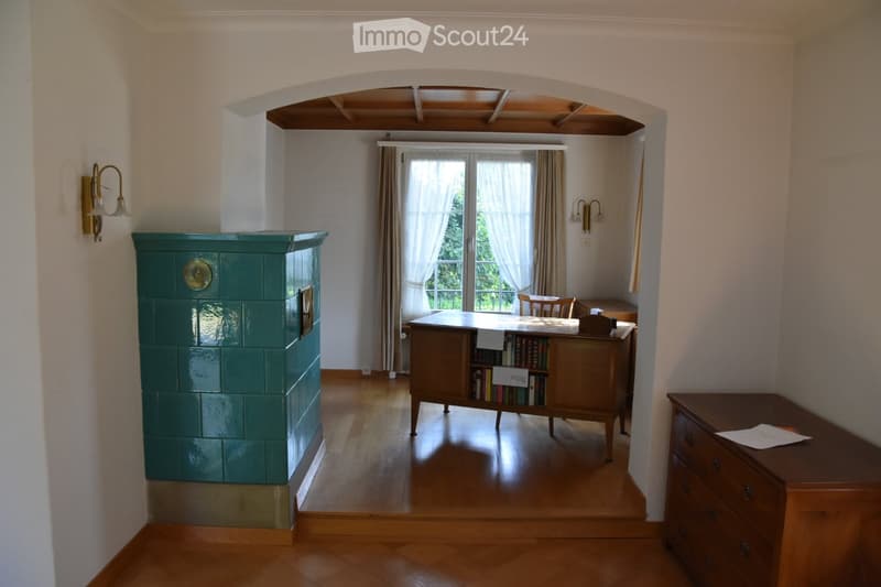 Brightly lit room with wooden flooring, a large window with curtains, a wooden desk with books, a cabinet, and a wall-mounted lamp.