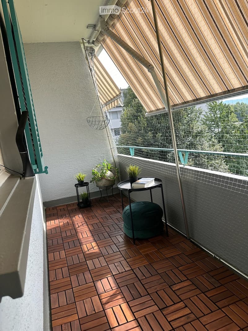 Balcony with wooden decking, glass railing, and striped awning. Includes a green stool, small table with potted plants and a book.