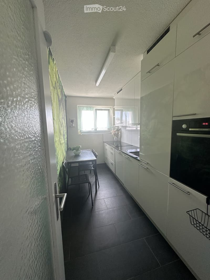 Modern kitchen with white cabinets, dark tiled floor, and a small dining area with a table and chairs. Features an oven, a vase with flowers, and a potted plant on the table.