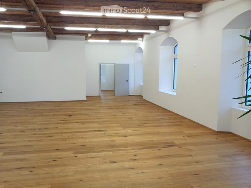 Empty room with wooden flooring, white walls, exposed wooden ceiling beams, and arched windows.