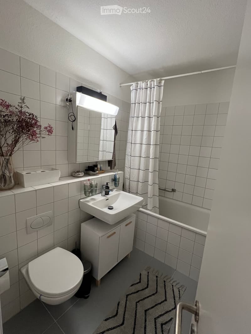 Modern bathroom with white tiles, a toilet, sink, mirror, bathtub, and shower curtain. Includes a vase with dried flowers on the counter.