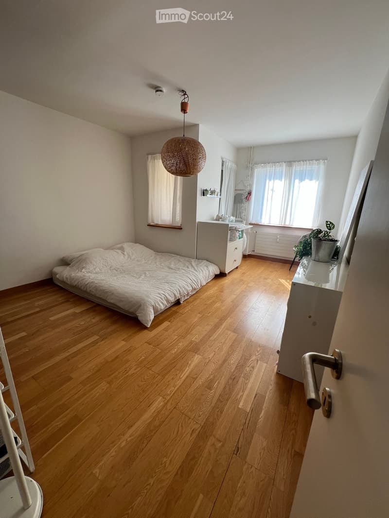 Cozy bedroom with wooden flooring, a single bed with white bedding, a nightstand with a mirror, a hanging woven lamp, and a small potted plant near the window.