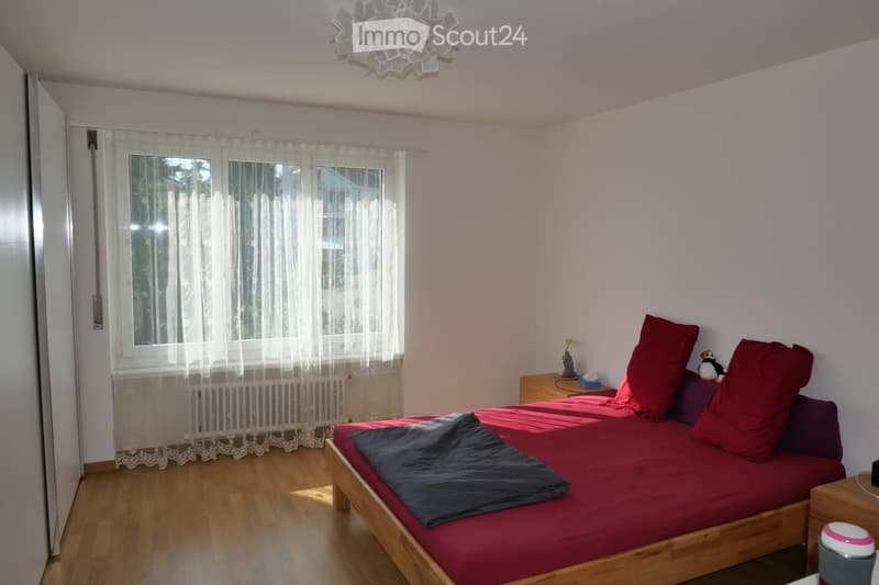 A cozy bedroom with a wooden bed, red bedding, a nightstand, and a window with sheer curtains.