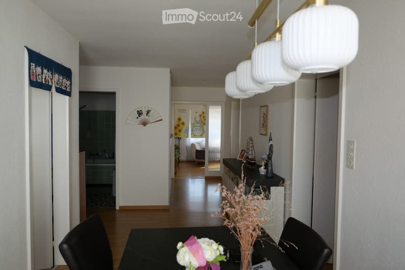 A modern hallway leading to a dining area with a black table, chairs, and decorative vases. The hallway features white walls, wooden flooring, and a hanging light fixture.
