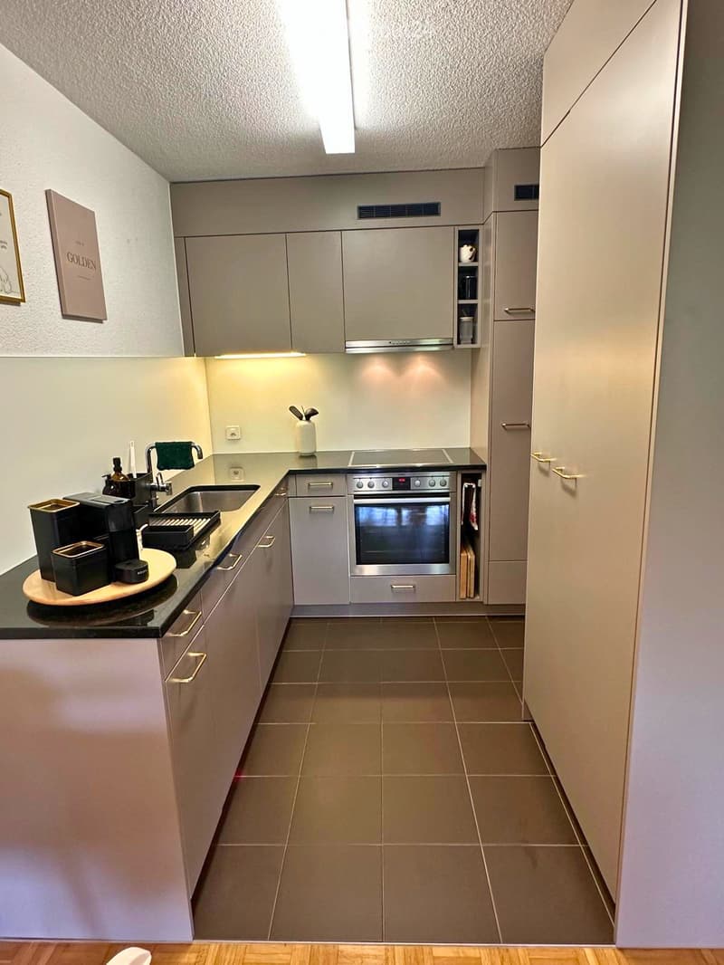 Modern kitchen with light grey cabinets, stainless steel appliances, and a black countertop. Features include a sink, oven, coffee maker, and various kitchen items.