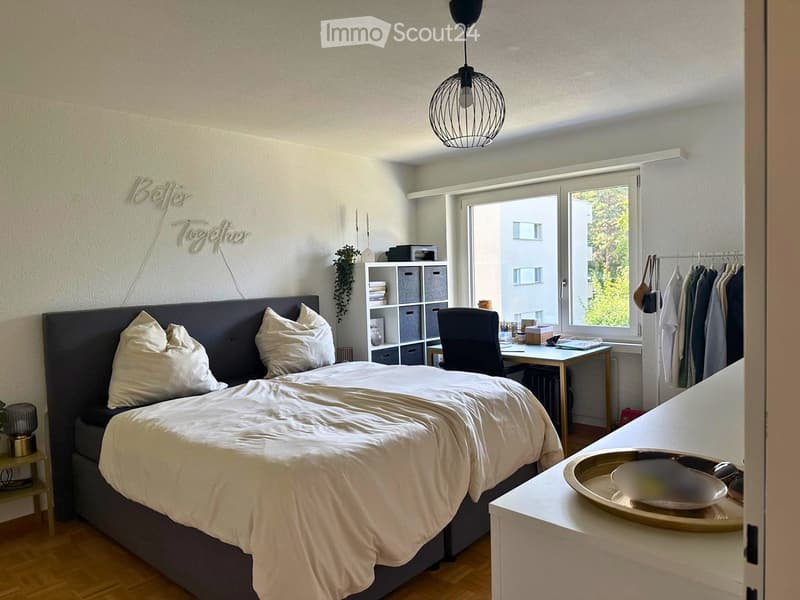 Modern bedroom with a large bed, white bedding, a desk with a chair, a shelf with storage boxes, and a hanging pendant light.