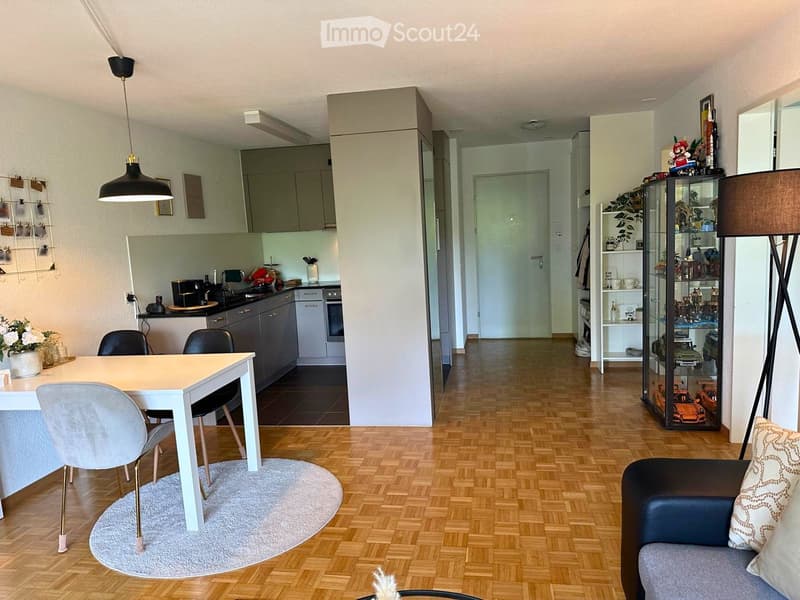 Modern open-plan living and dining area with wooden flooring, grey sofa, white dining table with chairs, kitchen area with cabinets and appliances, decorative shelf with figurines, and pendant lights.