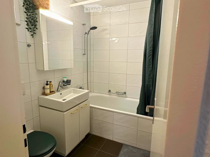 Modern bathroom with white tiles, a sink with a cabinet, a bathtub with a shower curtain, a toilet, and a mirror above the sink.