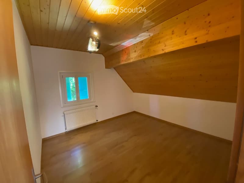 Empty room with wooden flooring, white walls, and a window with blue-tinted glass. The ceiling is wooden with exposed beams and a light fixture.