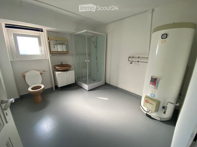 Modern bathroom with a toilet, sink, mirror, glass shower stall, and a water heater. The room features a grey floor and white walls.