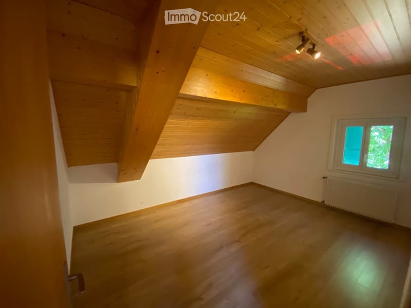Empty room with wooden floor and slanted wooden ceiling, featuring a window with a view of greenery and built-in lighting fixtures.