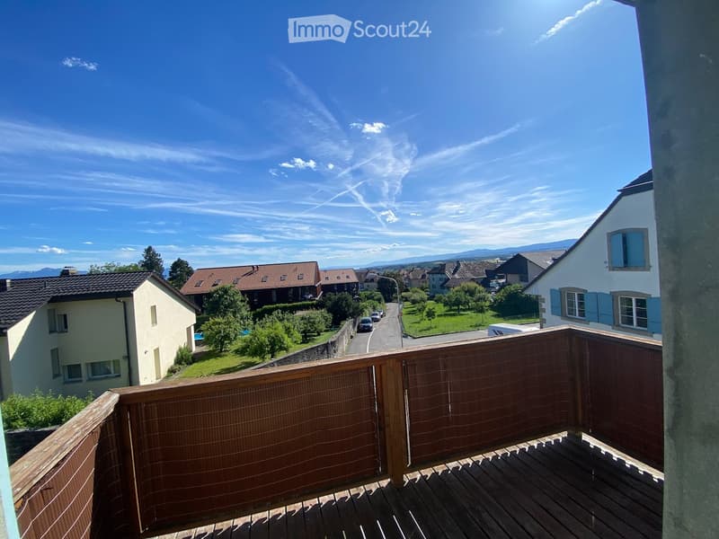 Balcony with wooden decking and railing, overlooking a residential neighborhood with houses, trees, and a clear blue sky.