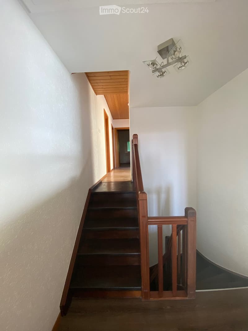 A narrow hallway with wooden stairs, white walls, and a wooden railing leading to an upper level. The ceiling features a wooden panel section and a modern light fixture.