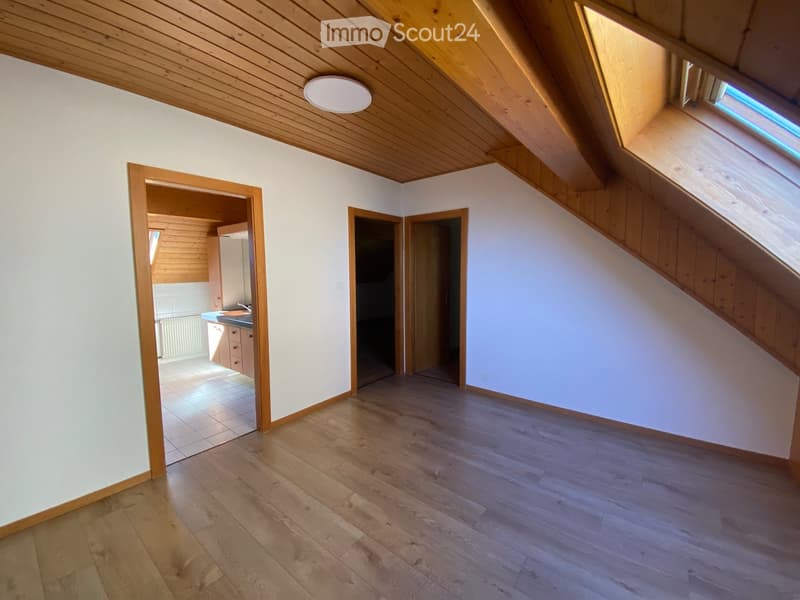 Empty attic room with wooden floors, white walls, and exposed wooden beams on the ceiling. Features multiple doors leading to other spaces, including a kitchen area visible in the background.