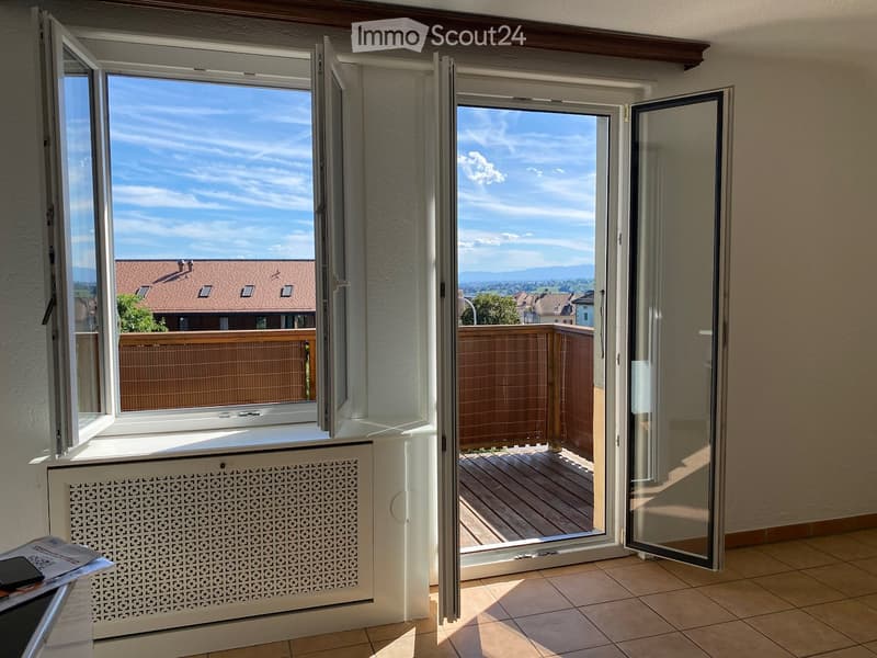 Bright room with large windows and sliding glass doors leading to a wooden balcony, offering a scenic view of the surrounding area.