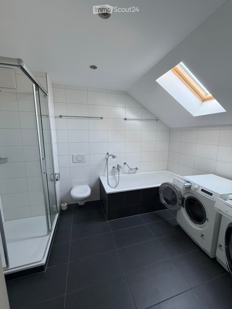 Modern bathroom with white tiles, dark grey floor, bathtub, toilet, shower, and washing machine. Features a skylight window and sleek fixtures.