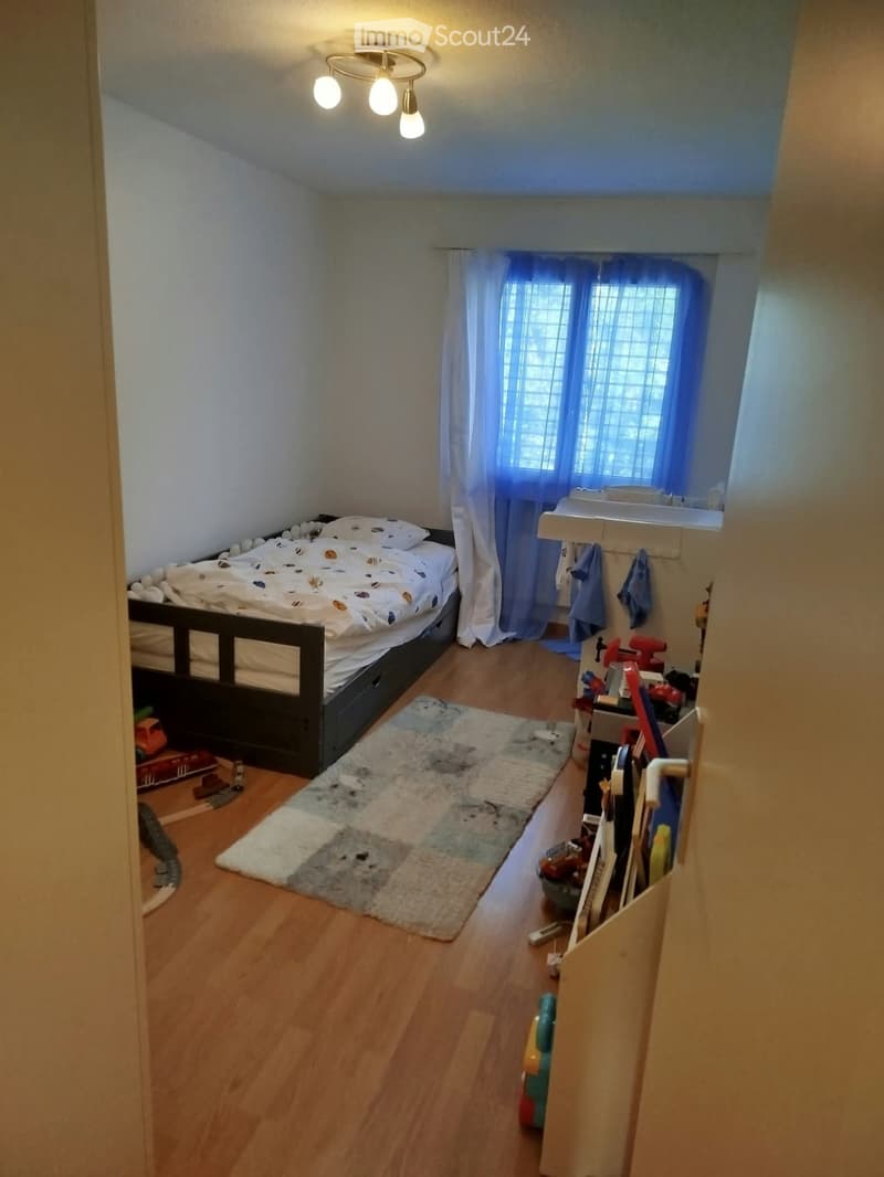 Cozy bedroom with a bed, patterned rug, small sink area, and toys visible. Wooden flooring and white walls.