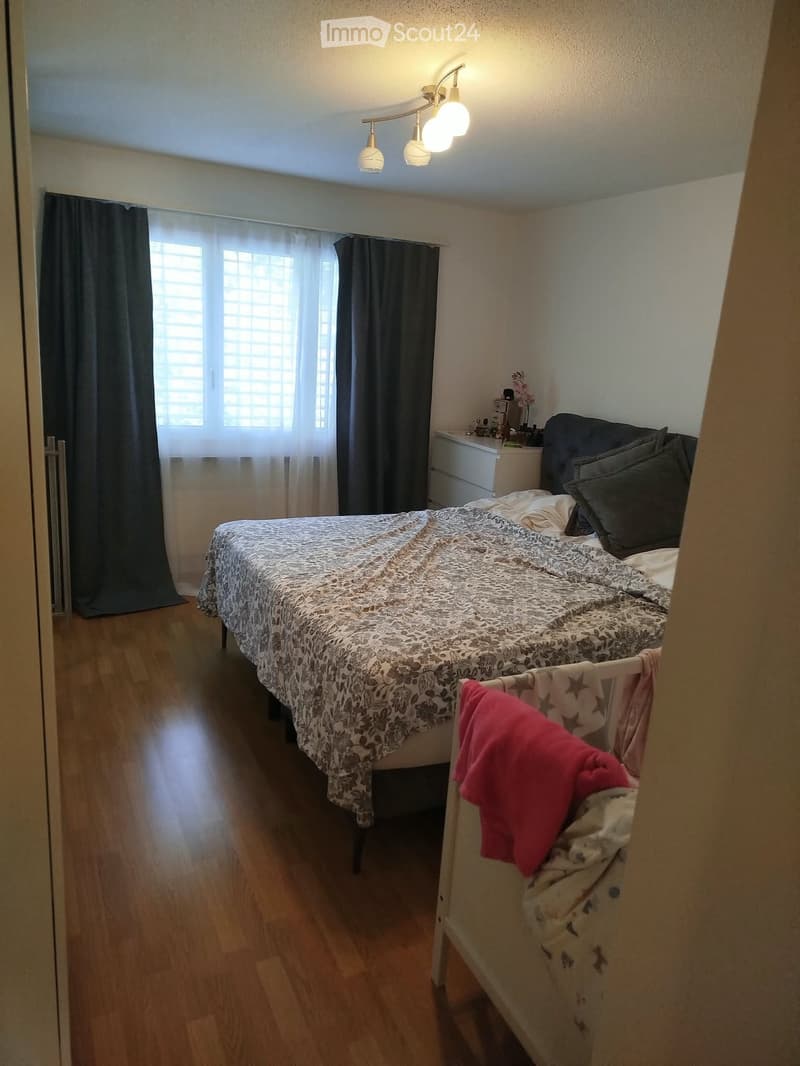 Cozy bedroom with wooden flooring, a double bed with patterned bedding, a nightstand, and a small baby crib with a red blanket.
