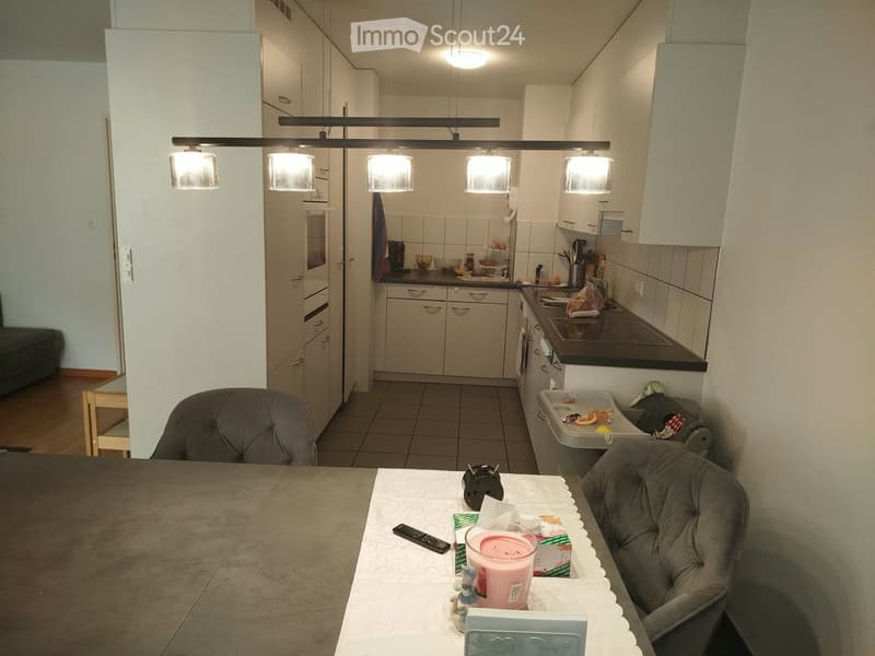 A modern kitchen and dining area with white cabinets, a grey dining table, two grey chairs, a candle, and a remote control on the table. The kitchen features a stove, sink, and refrigerator.