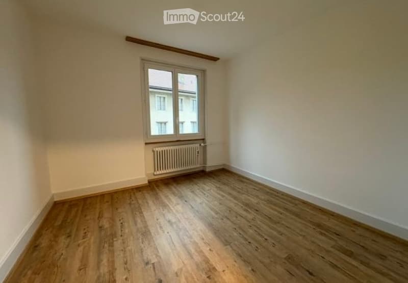 Empty room with wooden flooring, white walls, a single window, and a radiator below it.