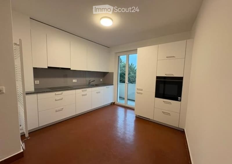 Modern, minimalist kitchen with white cabinets, a dark countertop, and a built-in oven. Features a large window with a sliding glass door leading to a balcony.