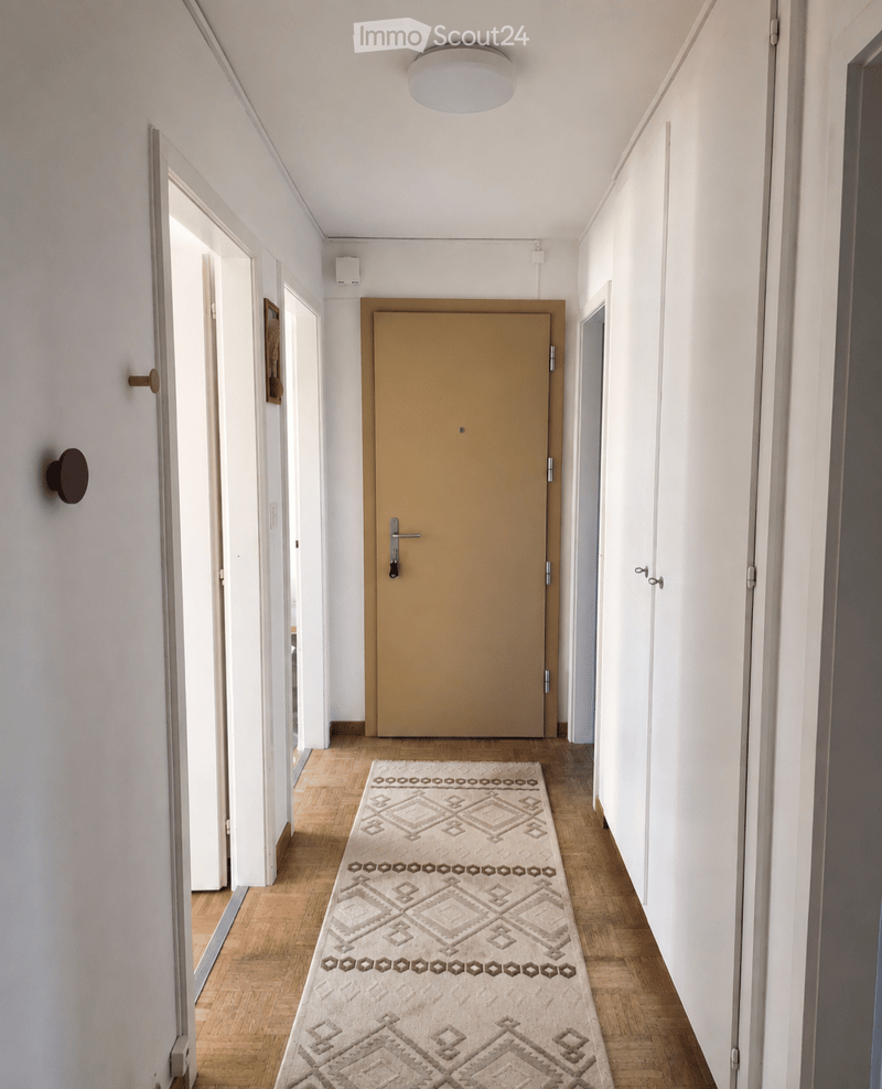 A modern hallway with light wooden flooring, white walls, and a patterned runner rug. Features a central closed door, several open doorways, and a ceiling light fixture.