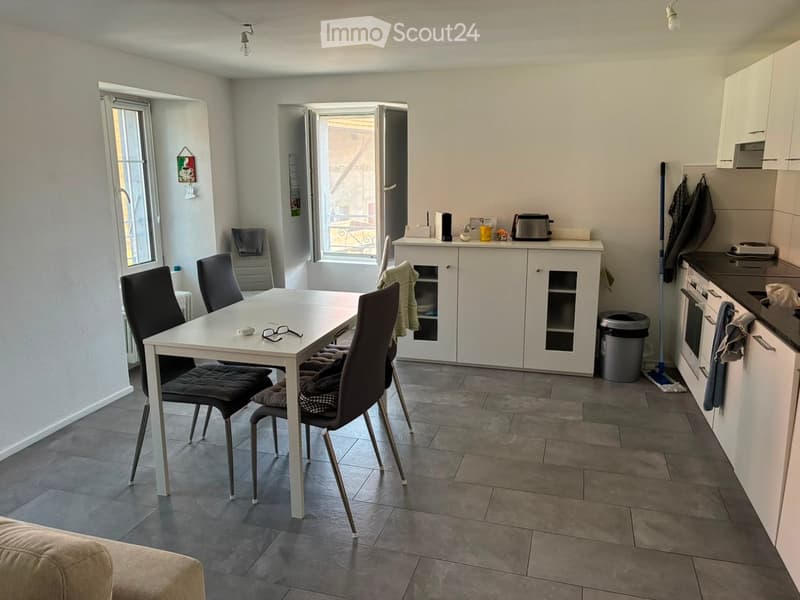 A modern, well-lit kitchen and dining area with a white dining table, four chairs, white cabinets, and a built-in oven. The space features tiled flooring and large windows that open to a balcony.