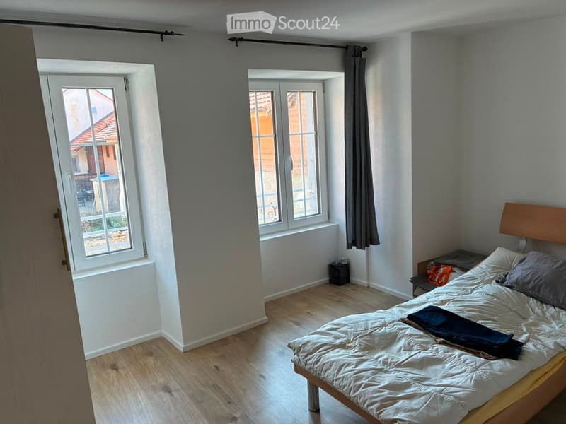 Bright bedroom with wooden flooring, large windows, a neatly made bed with pillows and a blanket, and a nightstand with a lamp.